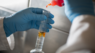 Close up image of a vial of clear liquid, two gloved hands can be seen holding the vial and adding drops of an orange  liquid into the vial.