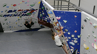 Image of students climbing on the NMT Bouldering Wall. The wall is made up of alternating blue and white panels.